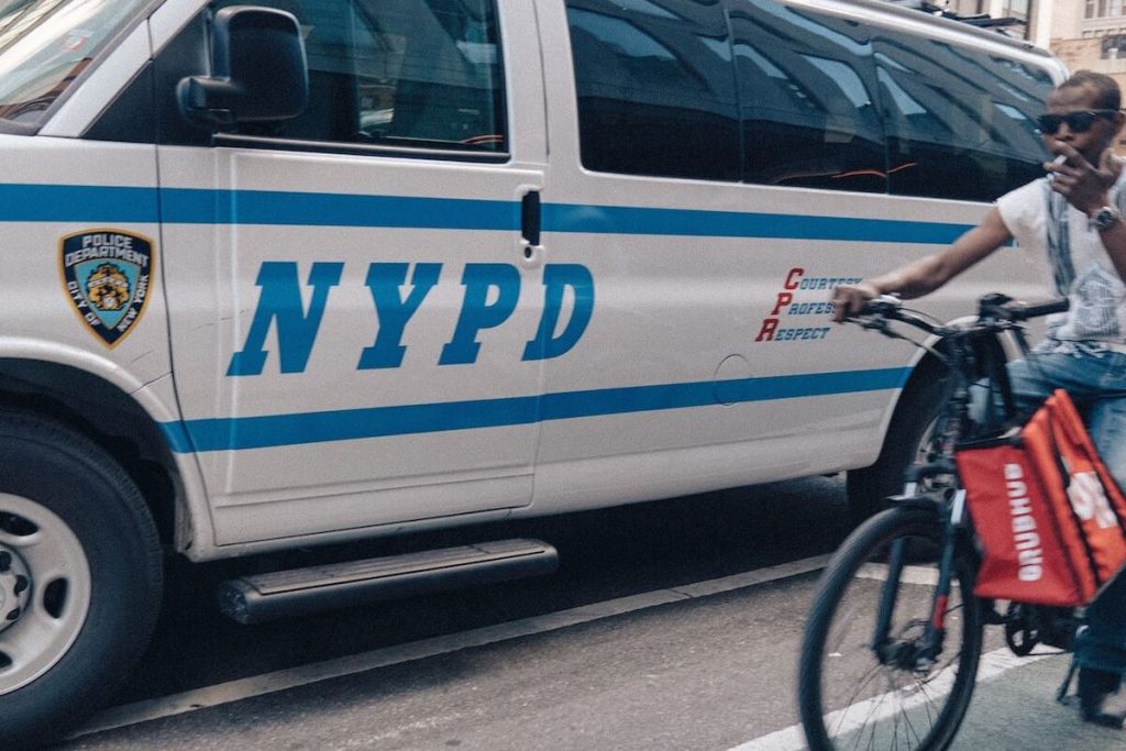 Image in a post titled 'nyc bike laws: 16 rules you need to know (2025 update)' | nypd van parked next to bike lane | fixed gear focus Food delivery cyclist breaking nyc bike laws next to police van.