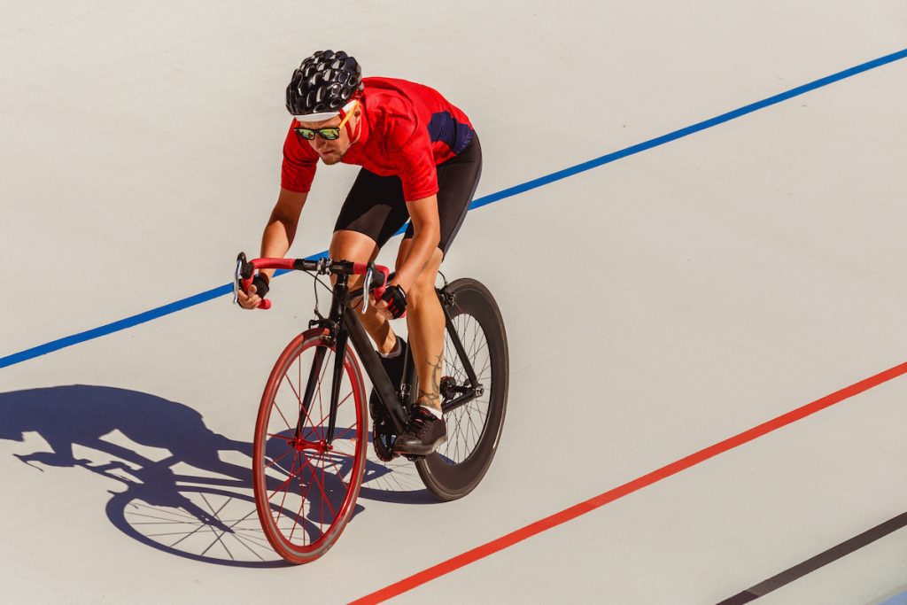 Image in a post titled 'global fixed-gear cycling events(that you can actually enter)' | image of a man on velodrome track wearing red jersey riding a track bike | fixed gear focus Image of a man on velodrome track wearing red jersey riding a track bike. Source: adobe stock